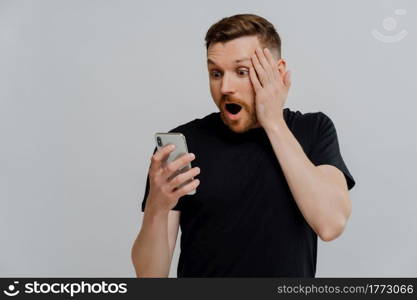 Young shocked impressed ginger man with stubble looking at mobile phone and reading stunning news, guy with red hair looking at smartphone in shock and touching face, isolated over grey background. Impressed man reading stunning news on smartphone with shocked expression