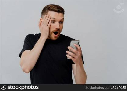 Young shocked impressed ginger man with stubble looking at mobile phone and reading stunning news, guy with red hair looking at smartphone in shock and touching face, isolated over grey background. Impressed man reading stunning news on smartphone with shocked expression