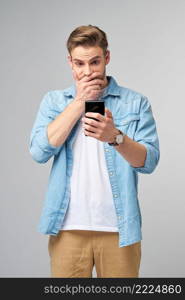 Young shocked stressed angry and frustrated man using smartphone over grey background.. Young shocked stressed angry and frustrated man using smartphone over grey background