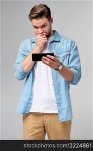 Young shocked stressed angry and frustrated man using smartphone over grey background.. Young shocked stressed angry and frustrated man using smartphone over grey background