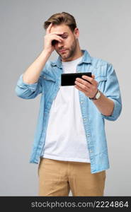 Young shocked stressed angry and frustrated man using smartphone over grey background.. Young shocked stressed angry and frustrated man using smartphone over grey background