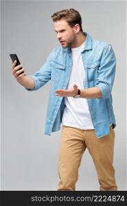 Young shocked stressed angry and frustrated man using smartphone over grey background.. Young shocked stressed angry and frustrated man using smartphone over grey background