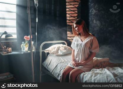 Young sick woman sitting on hospital bed on drip. Illness of female patient in clinic, health recovery and treatment. Young sick woman sitting on hospital bed on drip