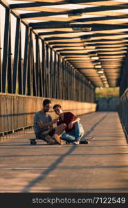 Young skateboarders sitting on their boards outside on a pedestrian bridge by the river, texting.