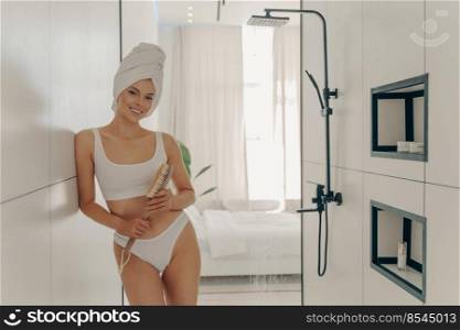 Young slender female model posing in modern light colored bathroom wears white sportive lingerie and wrapped towel on head, holding wooden brush for dry massage. Beauty treatment and body care concept. Young slender female model posing in modern light colored bathroom