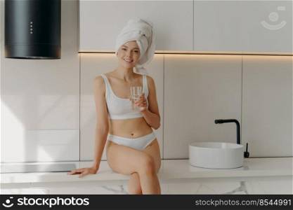 Young slender smiling thirsty woman holding glass of clean water and smiling at camera while sitting on white table top next to sink in stylish kitchen interior in morning, wears classic underwear. Young slender smiling thirsty woman holding glass of clean water and smiling at camera