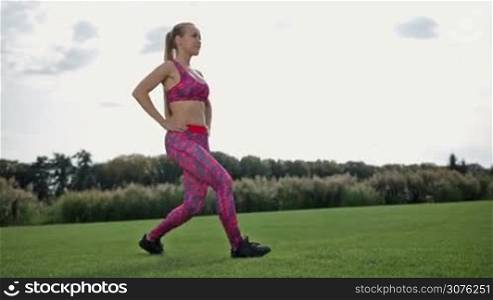 Young slim athletic woman doing frontal lunges and squats in the park. Athletic woman warming up before her morning workout outdoor on green grass. Side view.