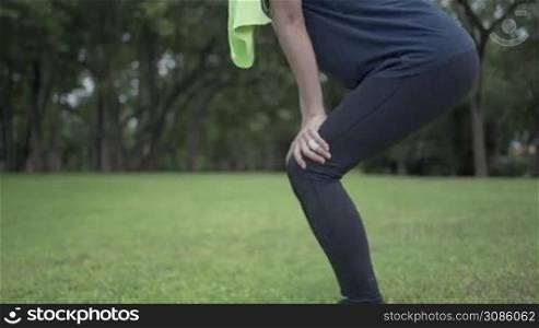 Young slim fit model in sportwear uses hands hold on knees while resting comfortable green meadow inside public park, injury from workout concept, morning time, people and healthy routine, body caring