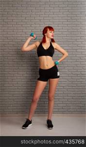 Young slim girl in sportswear is engaged in training and lifts dumbbells against a gray brick wall. girl lifts dumbbells
