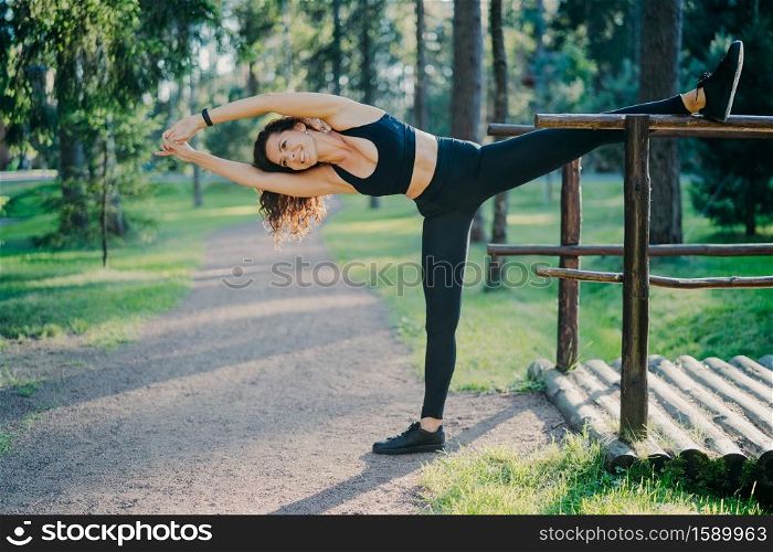 Young slim woman stretches outdoors, demonstrates her flexibility, wears cropped top and leggings, leans on left, poses outdoor in park, makes aerobic exercises. Active lifestyle and fitness concept