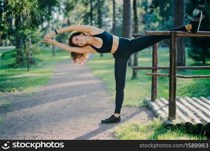 Young slim woman stretches outdoors, demonstrates her flexibility, wears cropped top and leggings, leans on left, poses outdoor in park, makes aerobic exercises. Active lifestyle and fitness concept