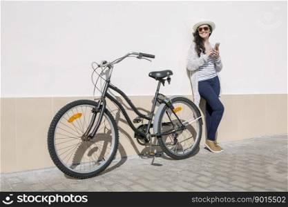 Young smiley woman in hat and sunglasses with bicycle and gadget standing against wall, Gran Canaria, Spain. . Smiley female with device and bicycle