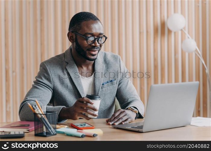 Young smiling african american business in glasses sitting with coffee cup at workplace in office, happy employee looking at computer screen with smile, reading internet news or checking email at work. Young smiling african american business in glasses sitting with coffee cup at workplace in office