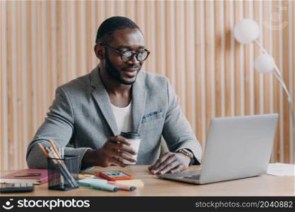 Young smiling african american business in glasses sitting with coffee cup at workplace in office, happy employee looking at computer screen with smile, reading internet news or checking email at work. Young smiling african american business in glasses sitting with coffee cup at workplace in office