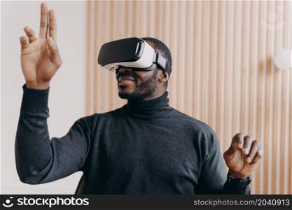 Young smiling african american businessman in VR glasses sitting in office, testing virtual reality goggles for business, impressed man using 3D technology, touching objects in air with hands. Smiling african american businessman in VR glasses sits in office, testing virtual reality goggles