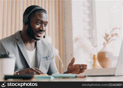 Young smiling african american man in headset consulting client customer via video call on laptop, pleasant dark-skinned guy foreign language tutor giving online lesson talking with students. Young smiling african american man in headset consulting client customer via video call on laptop