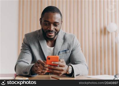 Young smiling african male entrepreneur sitting at office desk and using mobile phone at workplace, handsome successful afro american businessman reading online news or sending email on smartphone. Young smiling african male entrepreneur sitting at office desk and using mobile phone at workplace