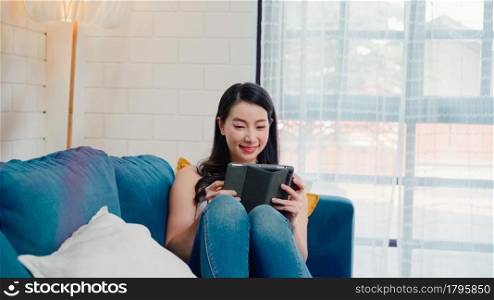 Young smiling Asian woman using tablet buying online shopping by credit card while lying on sofa when relax in living room at home. Lifestyle women at house concept.