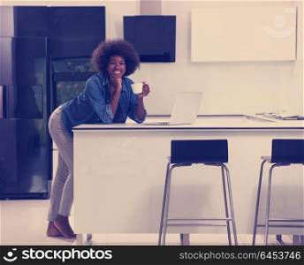 Young smiling black woman using computer and drinking coffee in modern kitchen interior