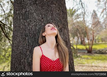 Young smiling blond woman with red dress leaning against a tree