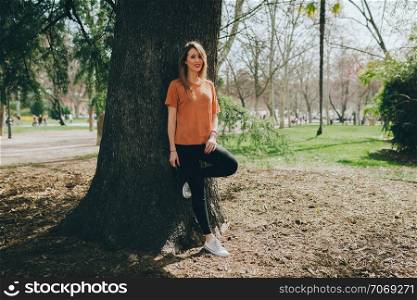 Young smiling blonde woman supported on a tree with black jeans