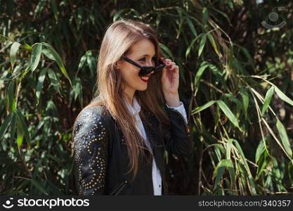 Young smiling blonde woman wearing a leather jacket in the park with sunglasses
