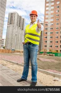 Young smiling construction engineer posing on building site with thumb up