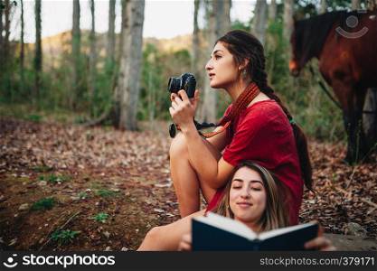 Young smiling couple of women wearing dresses reading a book and taking photos with old camera in the forest with their horse.