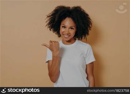 Young smiling dark skinned female curly hair pointing aside with thumb, shows blank space for promotion or advertisement with positive face expression, isolated over beige wall. Young smiling dark skinned female curly hair pointing aside with thumb, isolated over beige wall