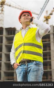 Young smiling engineer in hardhat and safety vest talking by phone on building site