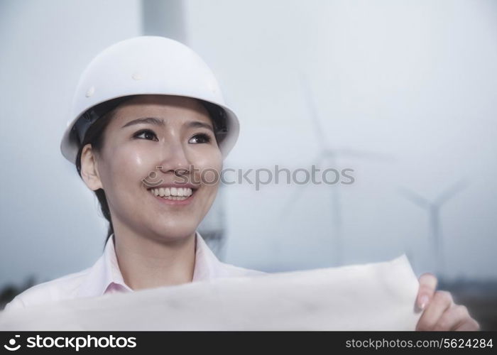 Young smiling female engineer holding a blueprint with wind turbines in the background