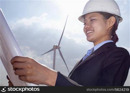 Young smiling female engineer holding open and looking down at blueprints, on site with wind turbines