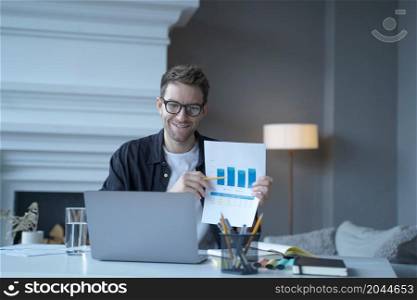 Young smiling german businessman in casual clothes pointing at document with graphics during internet meeting and talking with colleagues online while looking at laptop screen and working in office. Young smiling german businessman pointing at document with graphics during internet meeting