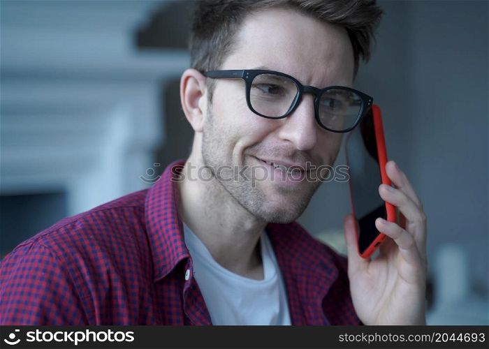Young smiling german guy in glasses using mobile phone and talking on with friend while spending leisure free time at home, happy man holding smartphone and making call to client during remote work. Young smiling german guy in glasses using mobile phone and talking on with friend
