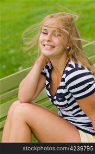 Young smiling girl sitting on the park chair and blowing wind mess up her hair