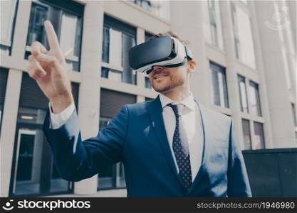 Young smiling handsome male enterpruer with stubble, dressed in formal attire, wearing portable VR goggles, trying to touch something in virtual reality with his finger, standing alone on city street. Cheerful businessman trying to touch something in virtual reality, standing near office building