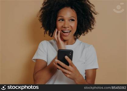 Young smiling happy Afro American woman using cellphone, overjoyed to see, to hear good news, isolated on beige background with copy space for advertising. Positive women emotions and body language. Young smiling happy Afro American woman using cellphone