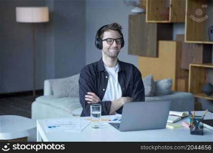 Young smiling happy austrian male freelancer sitting with closed eyes in headphones and enjoying listening to music at home office, man listens podcasts or audiobooks during remote work on laptop. Young happy austrian male freelancer in headphones enjoying listening to music at home office