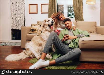 Young smiling heterosexual couple together on sofa with their dog using smartphone