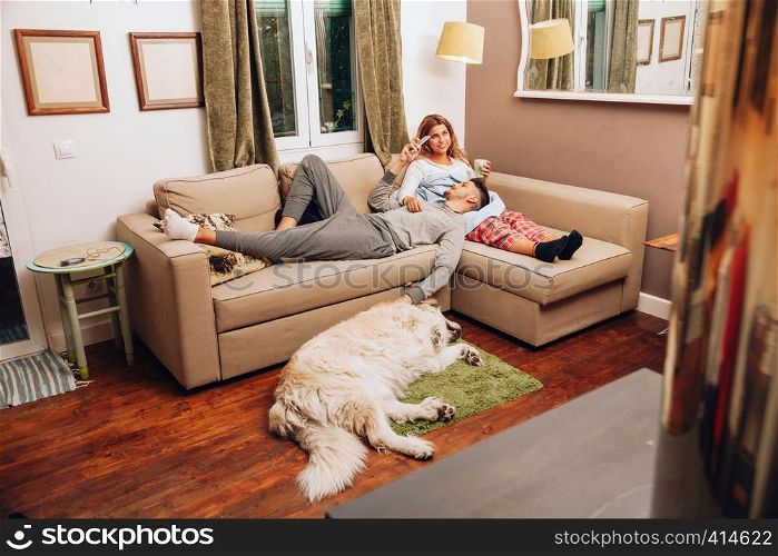 Young smiling heterosexual couple together on sofa with their dog using smartphone and drinking from a cup