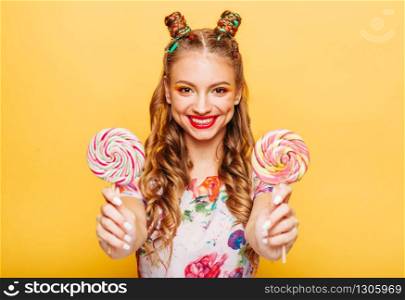 Young smiling lady holding two huge colorful lollypops. Amasing young woman fills happy and holding big candies in her hands. Stylish girl in summer dress, yellow wall on background.. Smiling lady holding two huge colorful lollypops