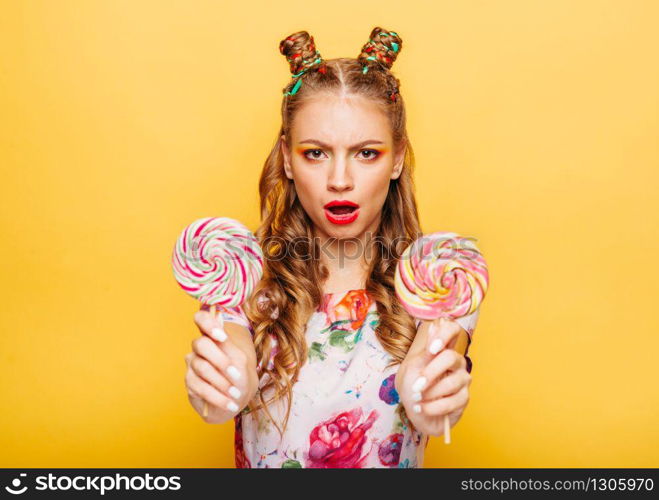 Young smiling lady holding two huge colorful lollypops. Amasing young woman fills happy and holding big candies in her hands. Stylish girl in summer dress, yellow wall on background.. Smiling lady holding two huge colorful lollypops