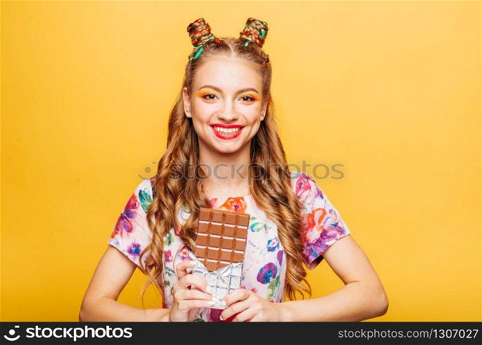 Young smiling lady holds chocolate bar. Amasing young woman fills happy and holding big chocolate in her hands. Stylish girl in summer dress, yellow wall on background.. Young smiling lady holds chocolate bar