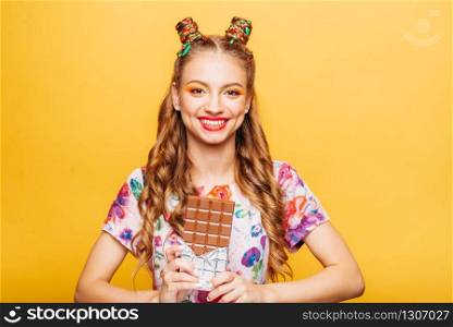 Young smiling lady holds chocolate bar. Amasing young woman fills happy and holding big chocolate in her hands. Stylish girl in summer dress, yellow wall on background.. Young smiling lady holds chocolate bar