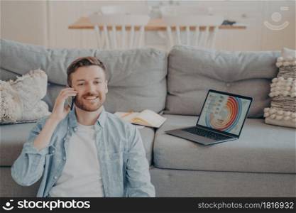 Young smiling male freelance worker calling his client to notify him about finished project, happy to receive payment, sitting on carpet while resting against couch, blurred background. Young smiling male freelance worker calling his client to notify him about finishing project