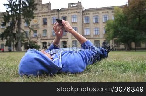 Young smiling male student in hoodie lying on the grass after classes outside univesity campus and browsing on the cellphone