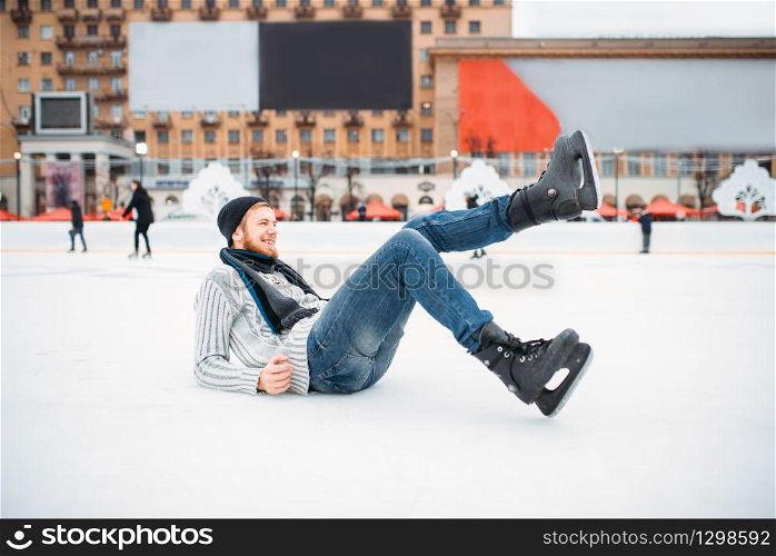 Young smiling man in skates sitting on ice, skating rink. Winter ice-skating on open air, active leisure. Young man in skates sitting on ice, skating rink