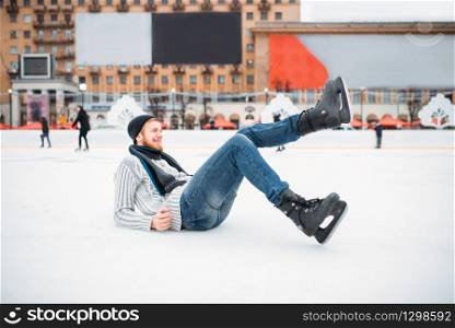 Young smiling man in skates sitting on ice, skating rink. Winter ice-skating on open air, active leisure. Young man in skates sitting on ice, skating rink