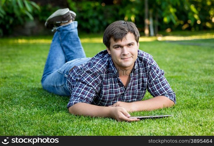 Young smiling man lying with tablet on lawn at park