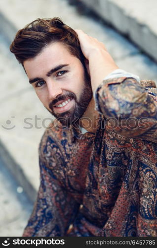 Young smiling man, model of fashion, wearing modern shirt in the street. Guy with beard and modern hairstyle in urban background.